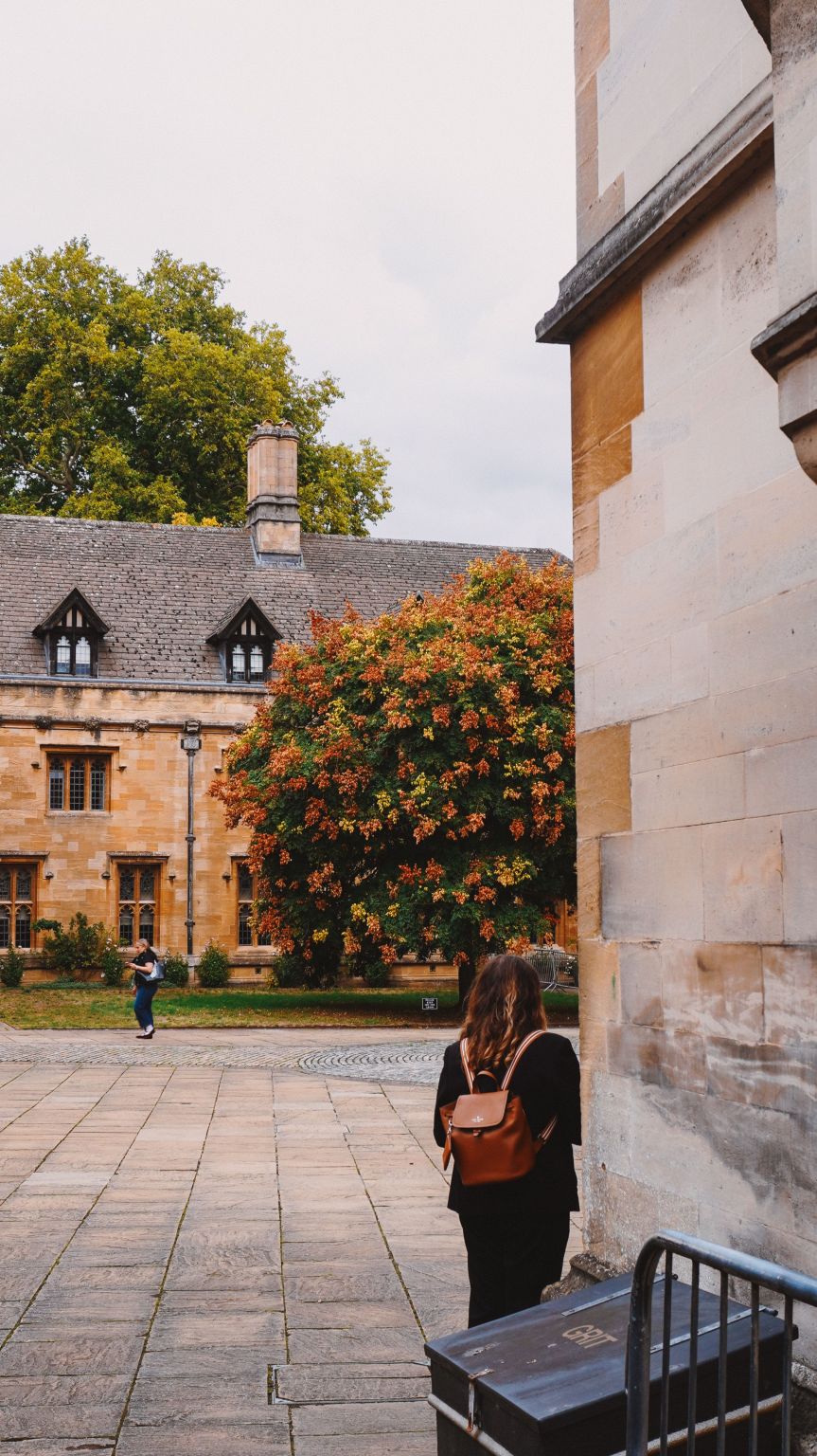 A student with a tan backpack in a paved courtyard, looking towards a stone college building and a tree in autumn colours